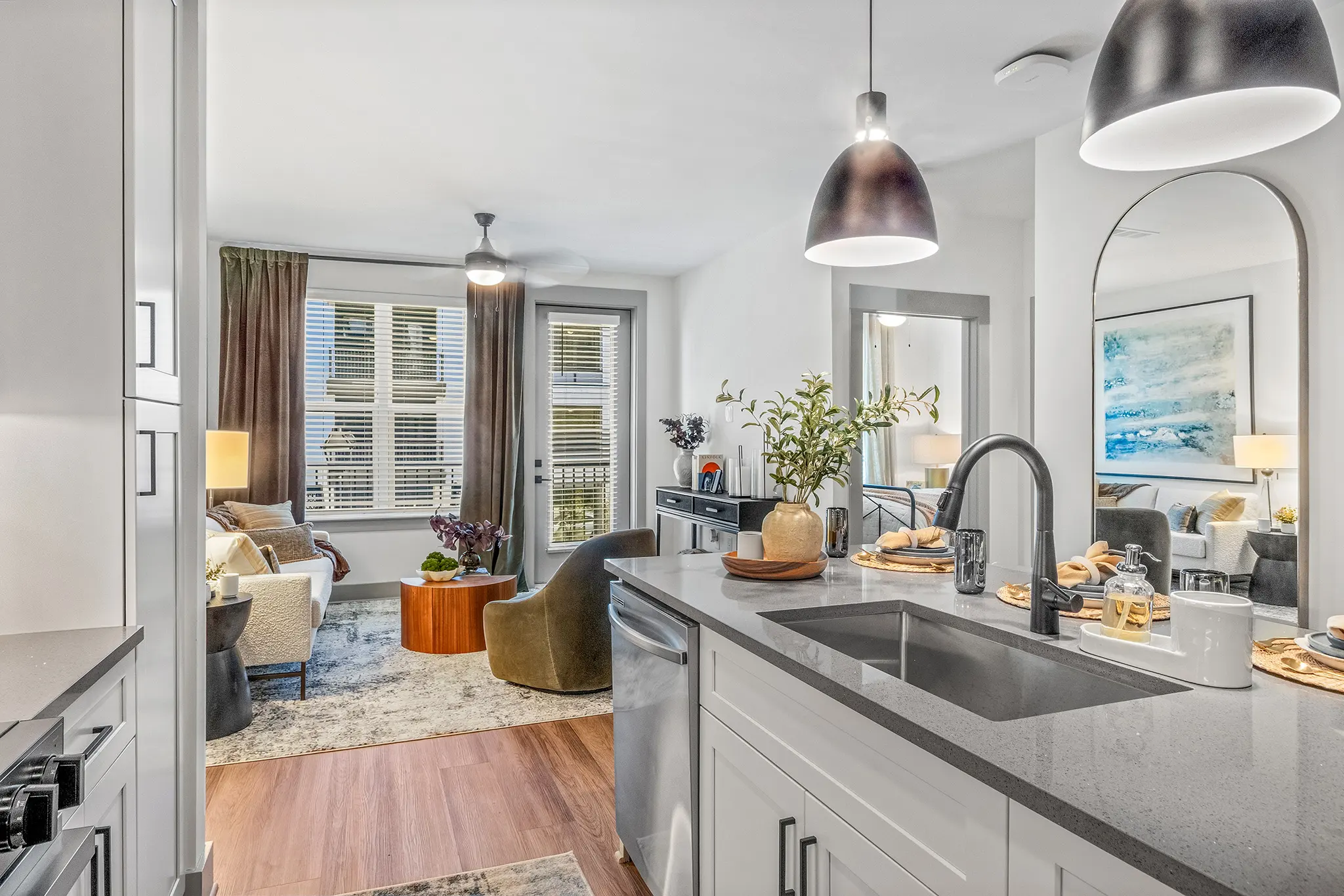 A modern apartment interior featuring a kitchen view into a cozy living area. The kitchen has white cabinetry and a dark countertop. Sunlight filters through large windows, illuminating the comfortable seating area with two armchairs and a sofa. A round coffee table sits on a light-colored rug, and decorative items adorn the space, enhancing its inviting atmosphere.