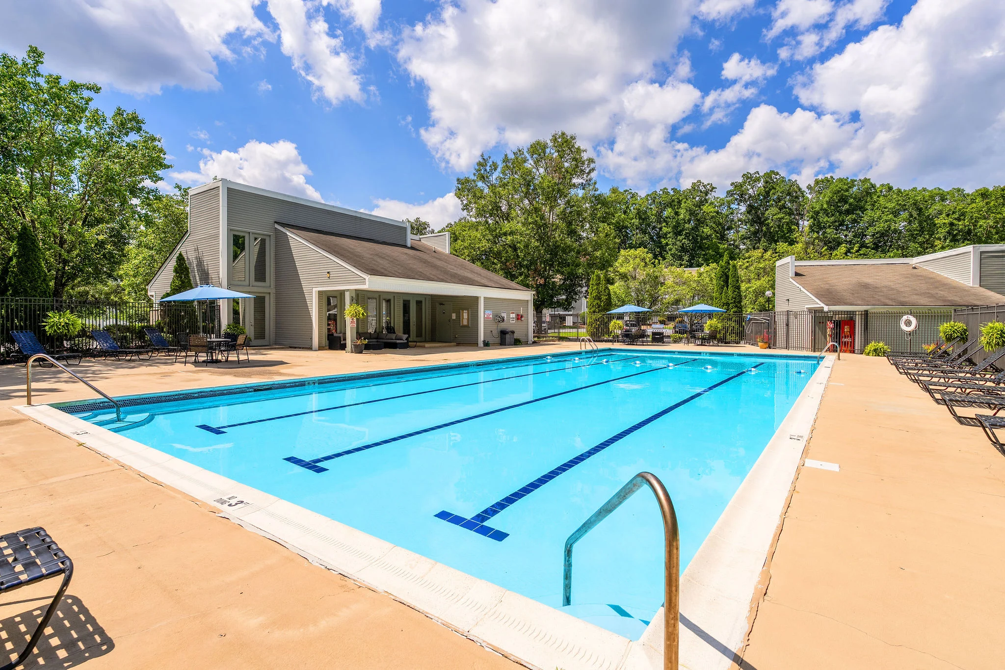 A clean, blue swimming pool surrounded by lounge chairs and green trees on a sunny day.