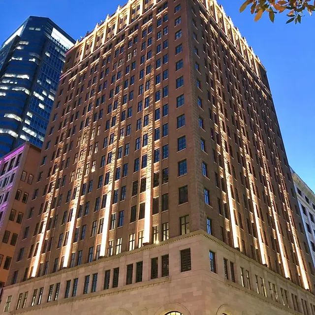 Historic Building Illuminated at Night A historic brick building with illuminated features against a twilight sky. The structure has multiple rows of windows and a prominent façade, showcasing architectural details. Trees partially frame the image, and modern buildings are visible in the background.
