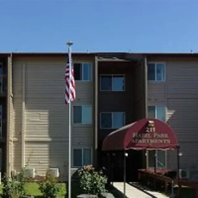Exterior view of Hazel Park Apartments, a three-story building with balconies, an American flag, and landscaped greenery.