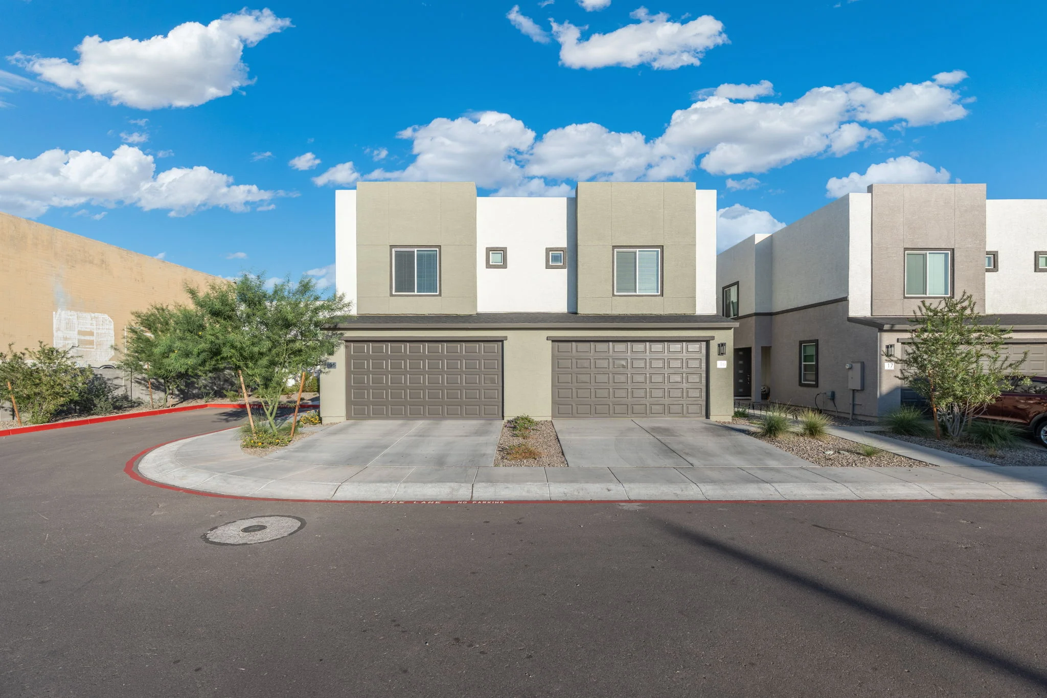 A modern two-story residential building with a gray and white facade, featuring two garage doors and surrounded by landscaping on a sunny day.