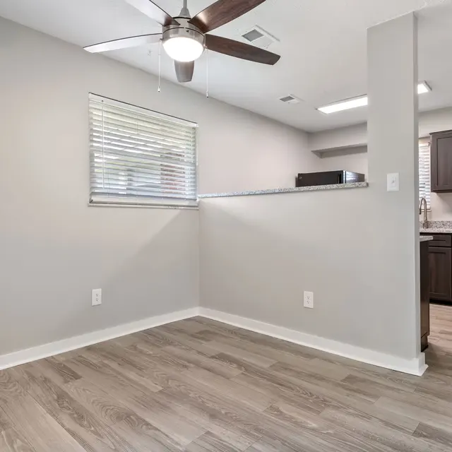 Modern, open living space with light-colored walls, wooden flooring, a ceiling fan, and a kitchen area with dark cabinets.