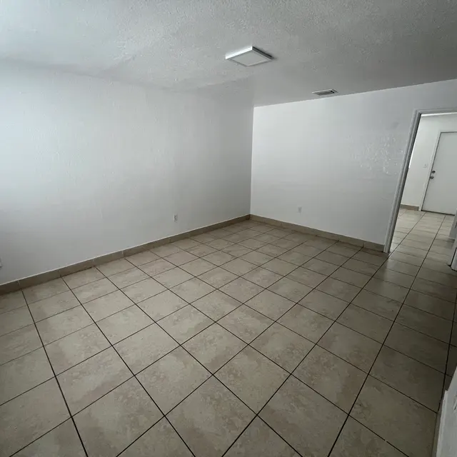 Empty room with white walls, tiled floor, and a door on the right; bright light from a ceiling fixture.