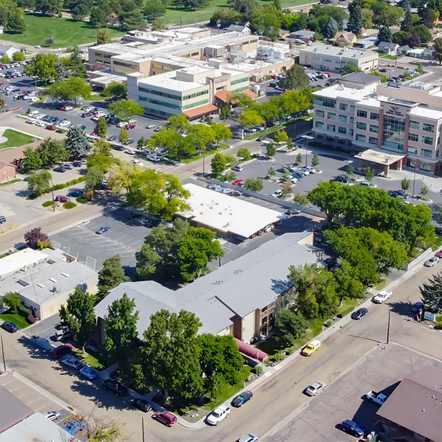 An aerial view of a town showing various buildings, parking lots, and greenery. There is a park area visible with trees and benches. The urban landscape includes a mix of commercial and residential structures.