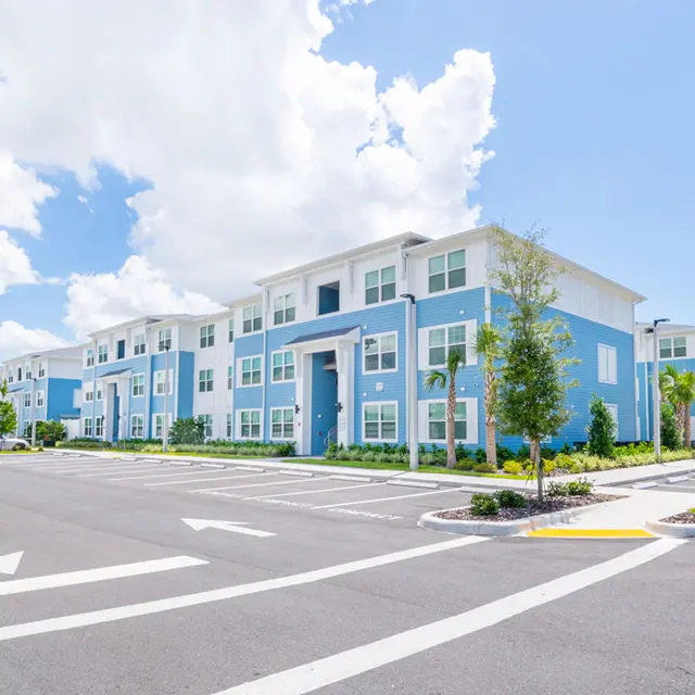 A modern apartment complex with blue and white exterior, featuring a spacious parking lot and landscaped areas under a bright blue sky with fluffy clouds.