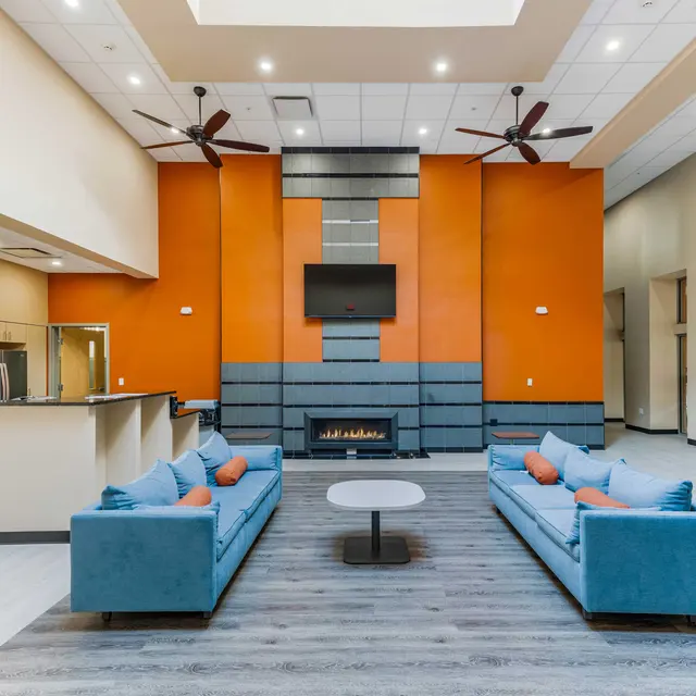 Spacious Modern Living Room A spacious modern living room with two blue sofas, a center table, an orange accent wall, and a fireplace. There are ceiling fans and a TV mounted on the wall.