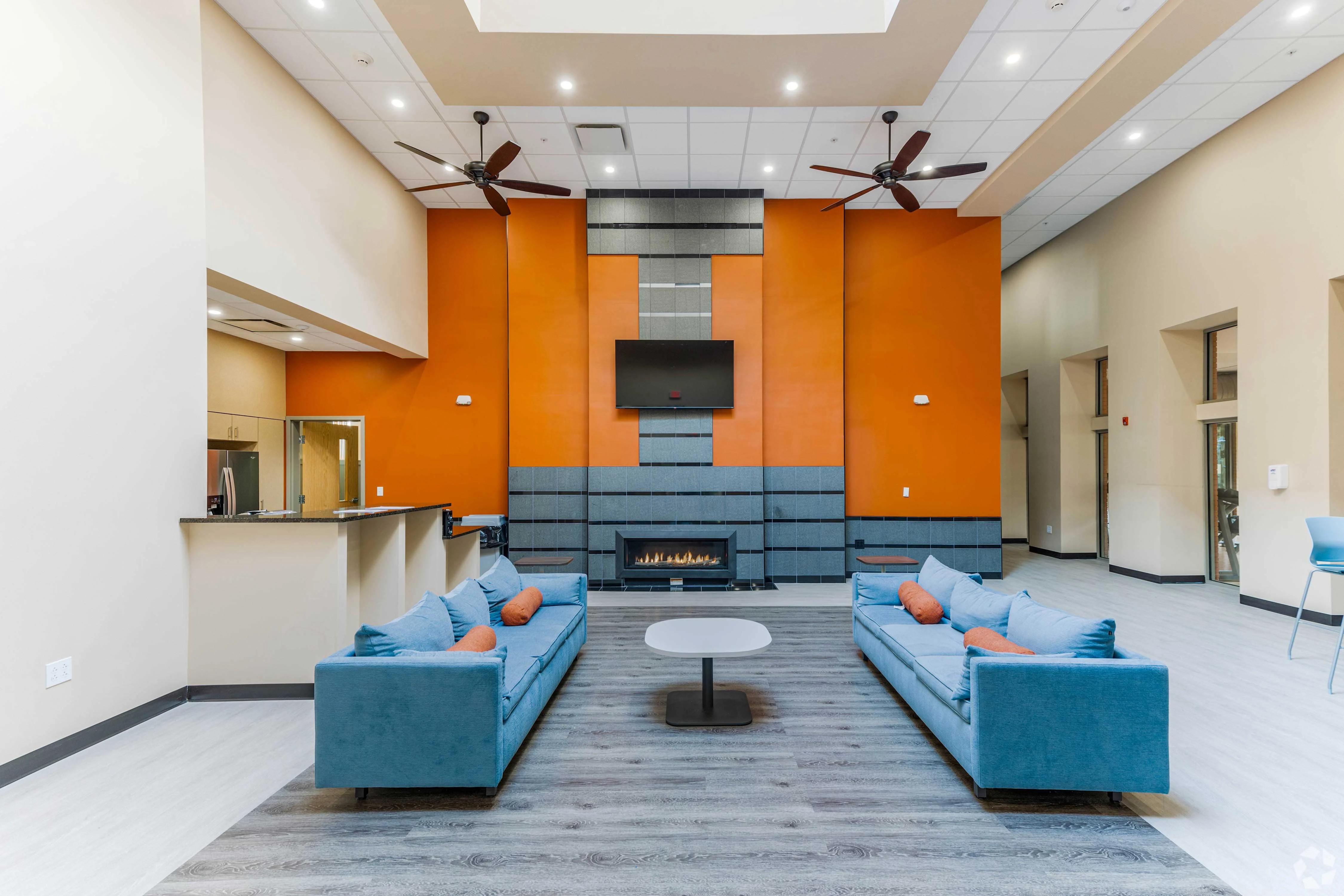 A spacious modern living room with two blue sofas, a center table, an orange accent wall, and a fireplace. There are ceiling fans and a TV mounted on the wall.