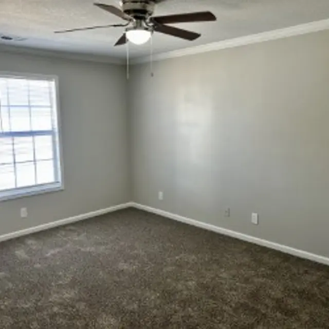 A spacious, empty room with beige walls and a ceiling fan. The floor is covered with a dark carpet. A window with white blinds is on the left side.