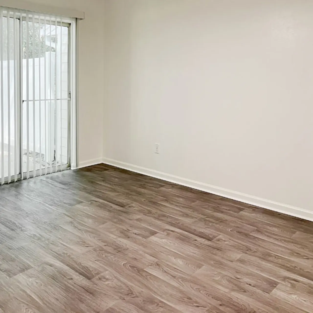 An empty room with light wood flooring and a window covered with vertical blinds.