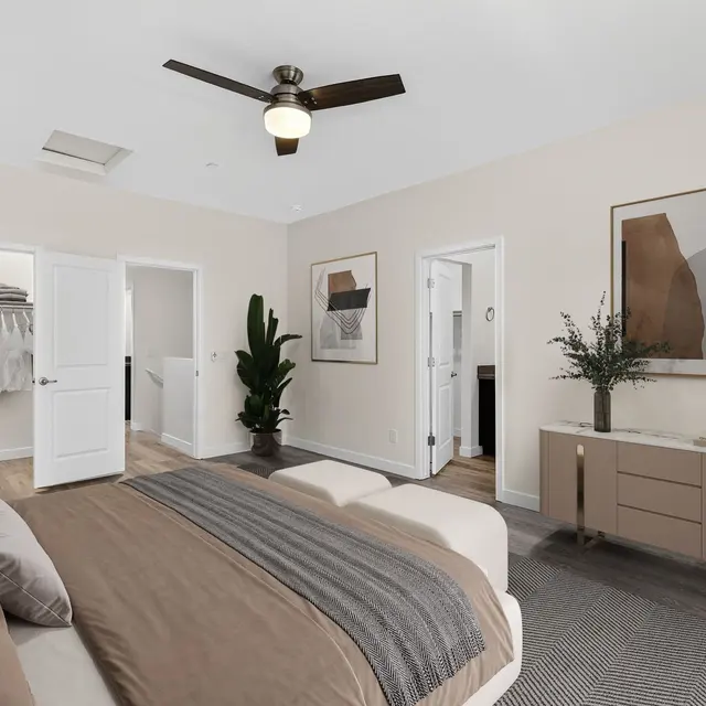 A modern bedroom featuring a large bed with neutral bedding, a ceiling fan, and two doors leading to separate areas. There's a closet with open doors displaying clothes, a green plant in the corner, and a wooden dresser with decorations on top.