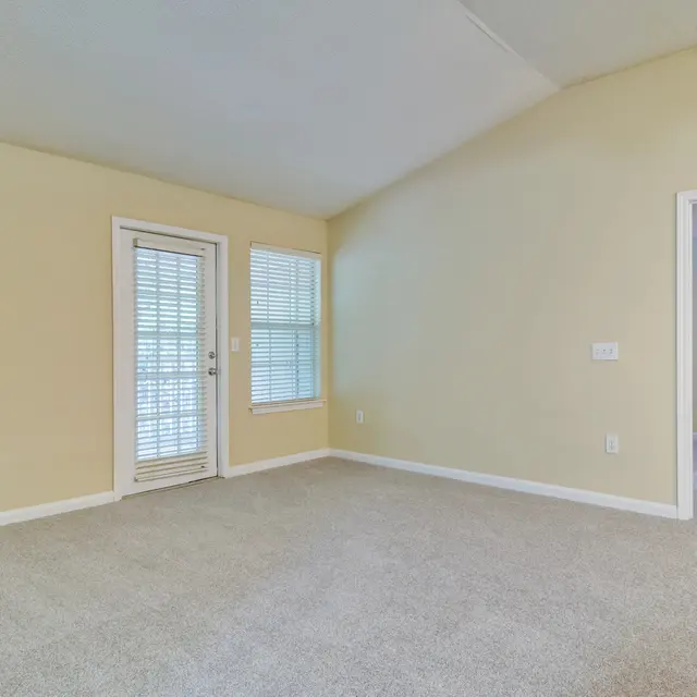 A spacious living room with light yellow walls, carpeted floor, and two sets of windows. One set of windows is a sliding door leading to an outdoor area, surrounded by additional windows with blinds. A doorway on the right leads to another room, and the ceiling is angled.