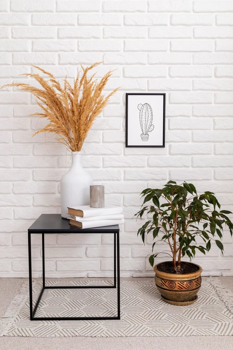 Minimalist Living Room Décor A minimalist living room corner featuring a black side table, a white vase with dried grass, and a small potted plant. A framed wall art depicting a cactus hangs on the white brick wall.
