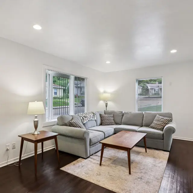 A cozy living room featuring a light gray sectional sofa, a wooden coffee table, and a lamp. The walls are white and there are two windows allowing natural light to enter the space. A rug lies on the dark wooden floor.