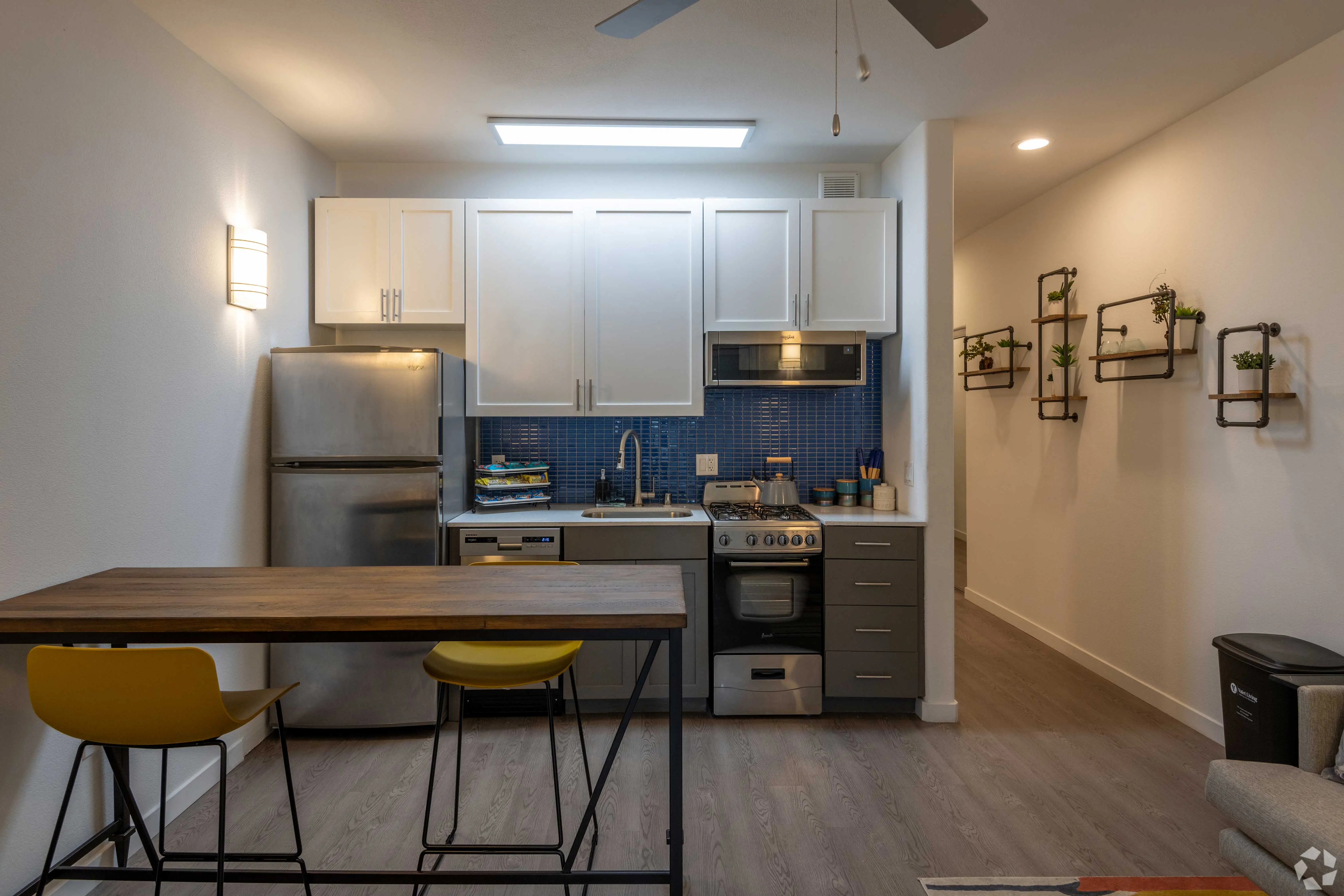 A modern kitchen featuring white cabinets, stainless steel appliances, and a blue tile backsplash. A wooden table with yellow stools is in the foreground, and there are wall-mounted planters along the adjacent wall.