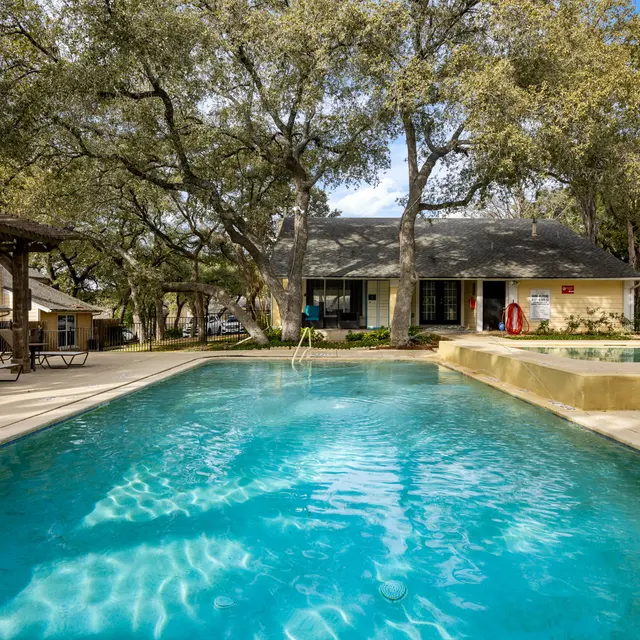 A view of a swimming pool surrounded by trees, with lounge chairs on one side and a building in the background.