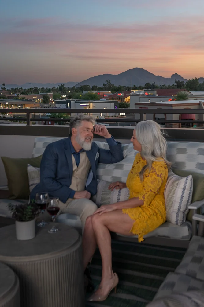 A man and woman sitting on a rooftop during sunset, enjoying a conversation. The man has a beard and is wearing a blue suit with a light vest. The woman is wearing a vibrant yellow dress with lace details. They are seated on a comfortable couch with a table in front of them, and there are glasses of red wine on the table. The background shows a sunset over mountains and city lights.