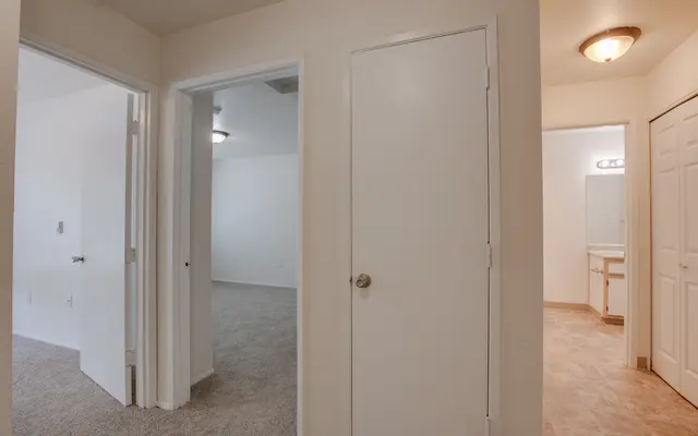 A view of a hallway with multiple doors leading to different rooms, including a bathroom and another room. The flooring is carpeted, and the walls are painted white.
