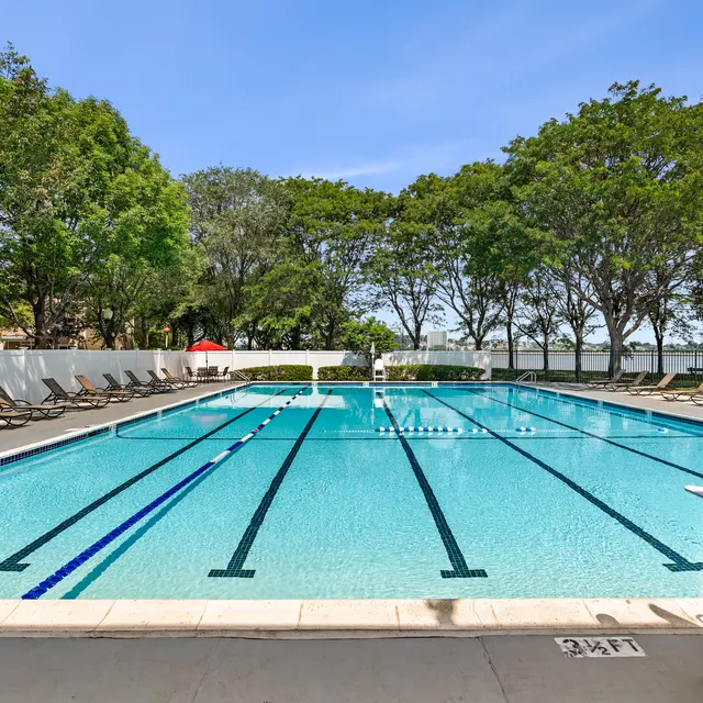A clean outdoor swimming pool with clear blue water, lane markings, and empty lounge chairs lining the edge. Trees in the background provide shade under a clear blue sky.