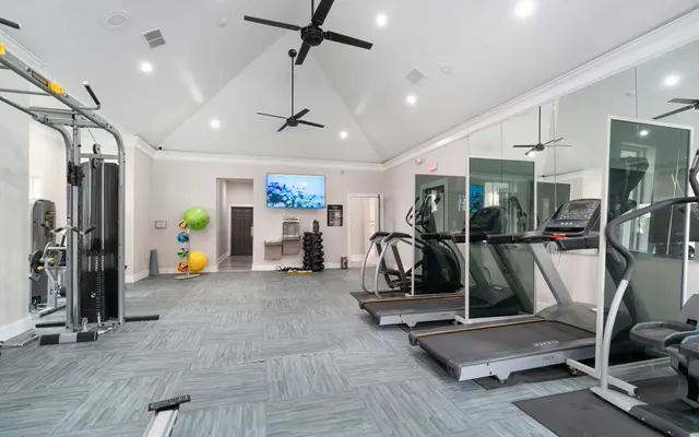 A fitness center with high ceiling fans, vinyl flooring, TV on the wall and cardio equipment. 