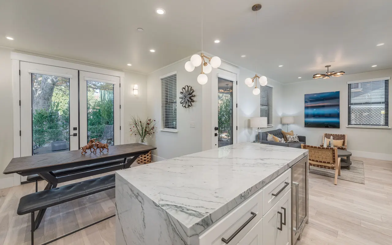 A modern dining area featuring a marble countertop island, a long wooden dining table with benches, and a cozy sitting area with a sofa and decorative art. Large windows allow natural light, and greenery is visible outside.