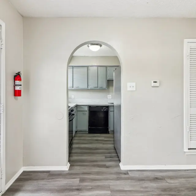 Interior view of a modern apartment featuring a small archway leading to the kitchen area, with light-colored walls and flooring, and large windows with blinds.