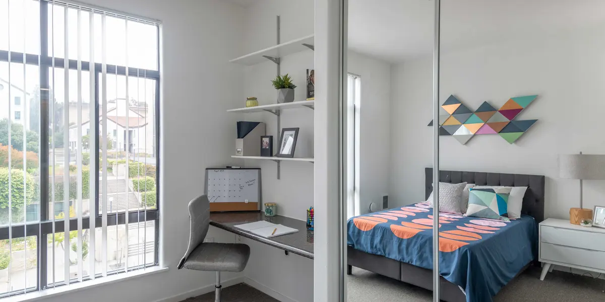 A modern bedroom featuring a neatly made bed with colorful bedding, a small desk and chair setup, and large windows allowing natural light to enter.