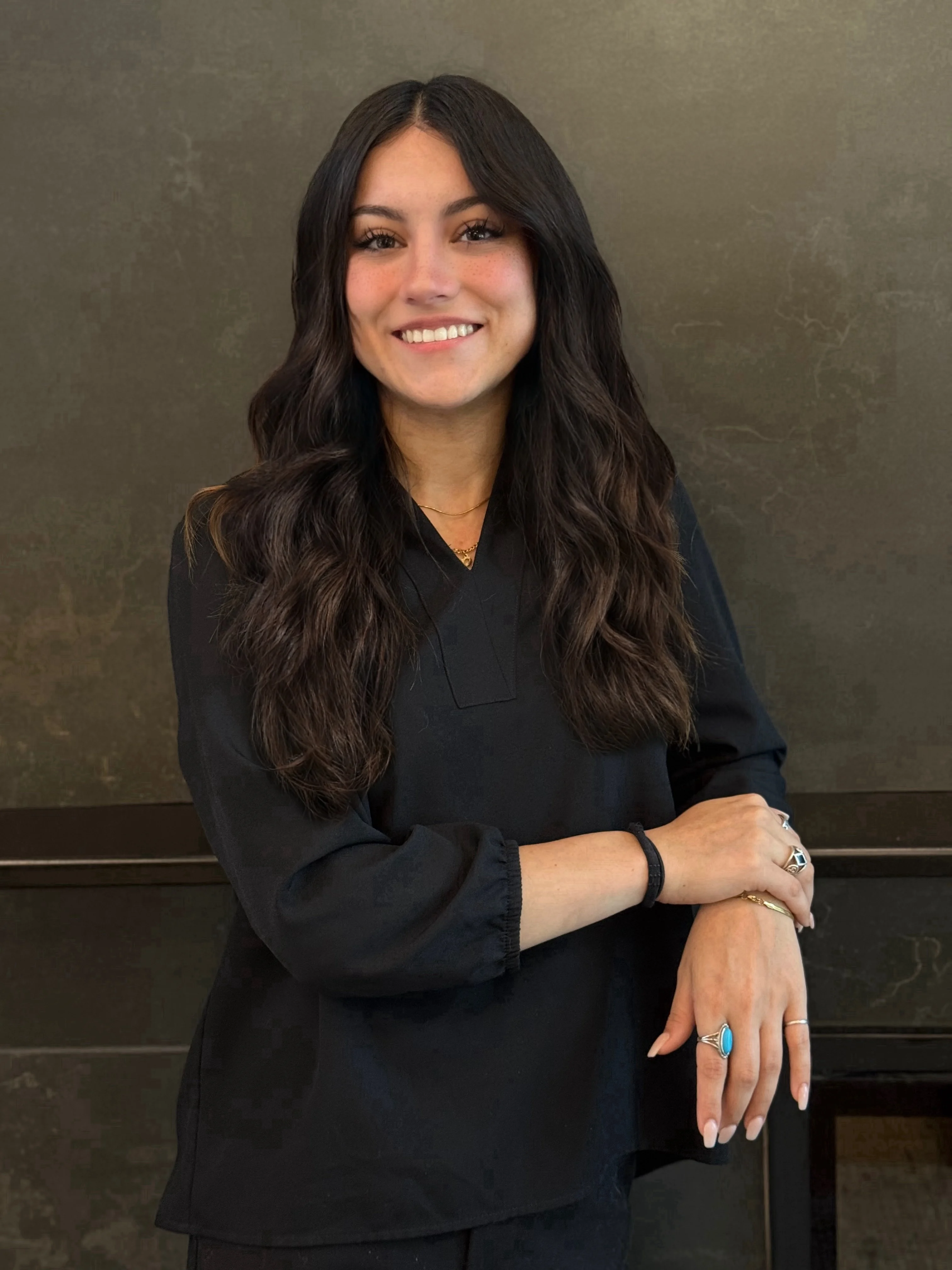 A woman with long dark hair, wearing a black top, smiling while standing with her arm crossed in front of a dark textured background.