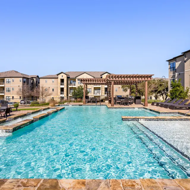 A large swimming pool at an apartment complex surrounded by lounge chairs and buildings in the background.