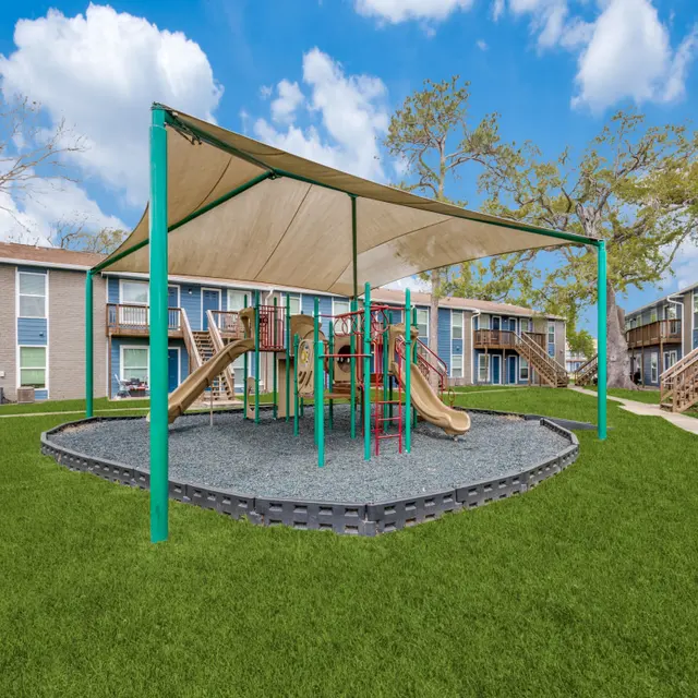A playground area featuring slides and climbing structures, covered by a large shade canopy, surrounded by green grass and apartment buildings in the background.