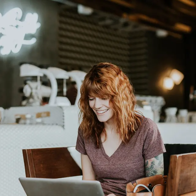 High Street Lofts Smiling woman with curly red hair working on a laptop in a cozy café with a lively atmosphere.