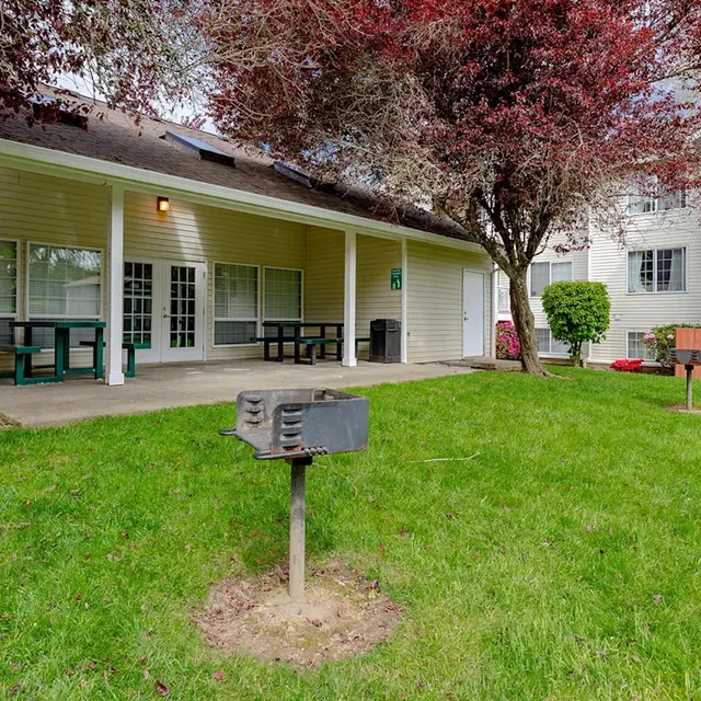 View of a backyard area with a patio and picnic tables, surrounded by lush green grass and trees, adjacent to a residential apartment building.