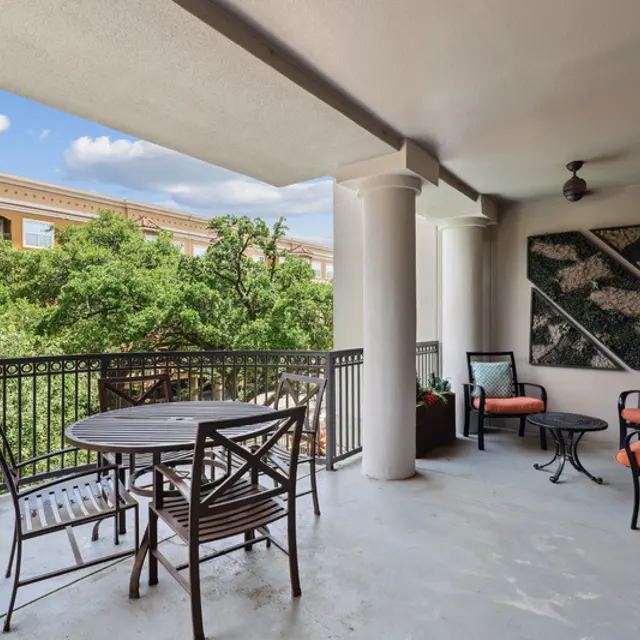A spacious balcony featuring a round table with four chairs, two armchairs, and a beautiful view of trees and a building in the background.