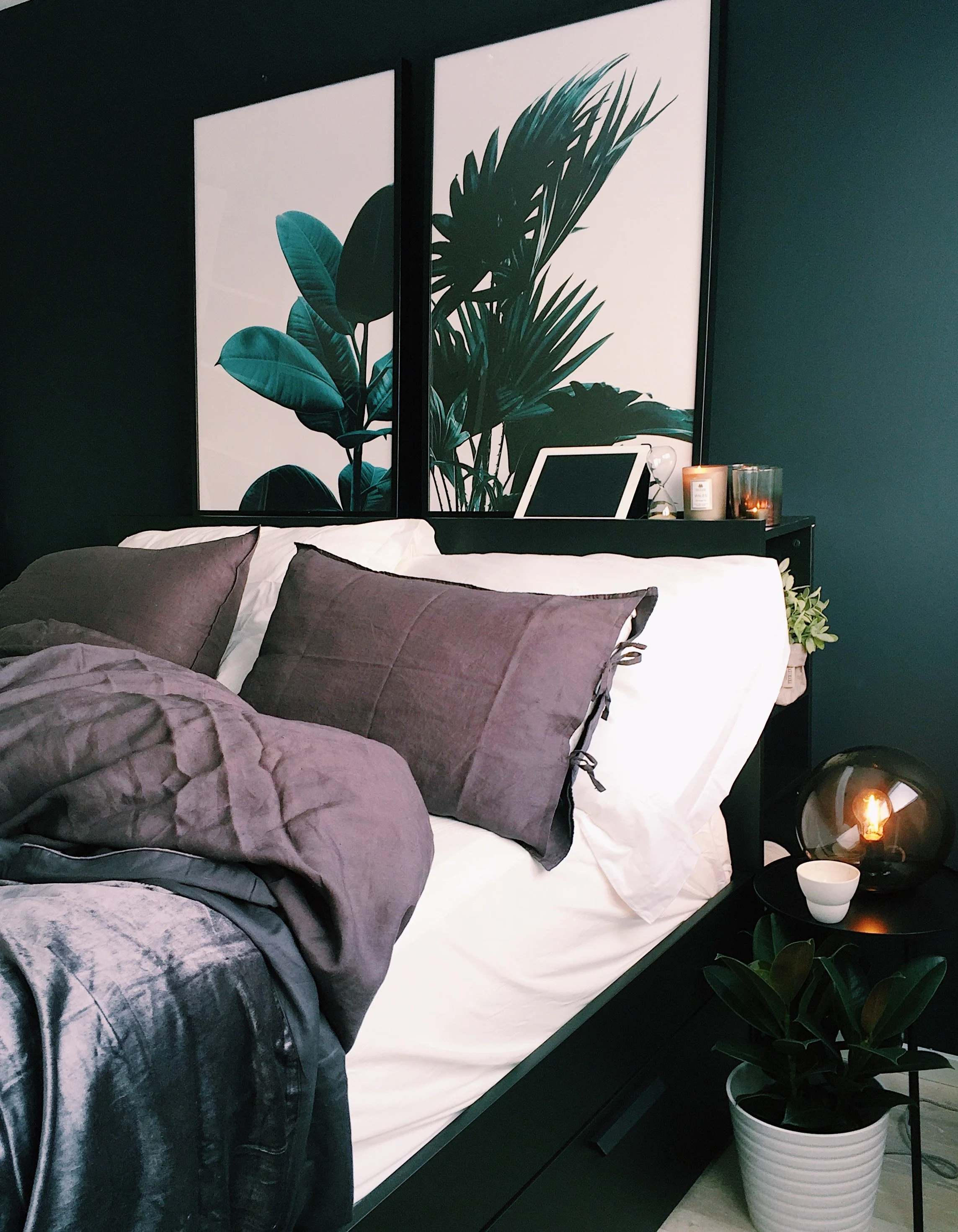 A cozy bedroom featuring a dark green wall, a neatly made bed with gray pillows and a soft blanket, decorative plants, and two framed leaf prints hung above the bed.
