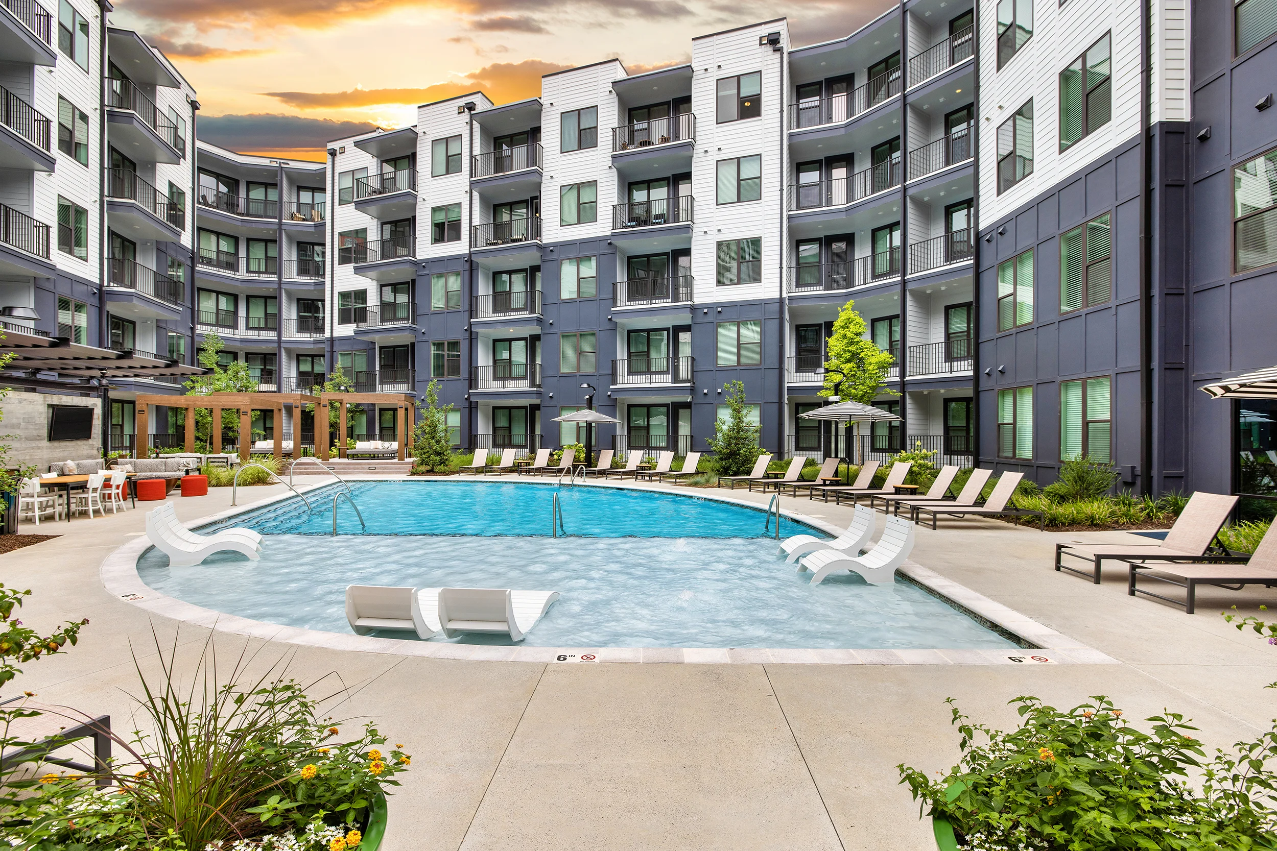 Modern Apartment Pool Area A modern pool area surrounded by apartment buildings. The pool is shaped in a circular design with lounge chairs around it, set against a backdrop of greenery and a sunset sky.