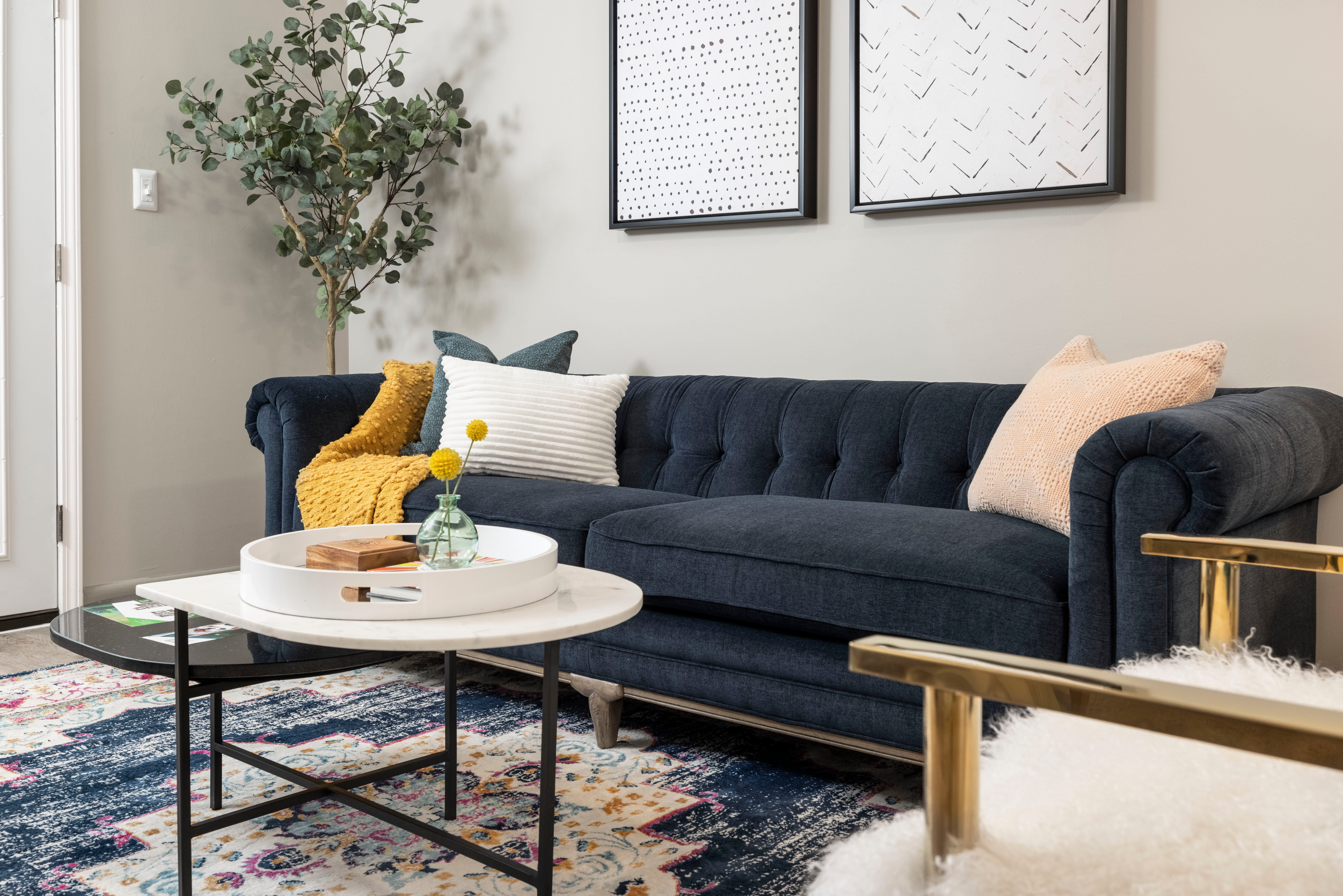 A stylish modern living room featuring a dark blue tufted sofa, a white round coffee table, patterned rug, and decor elements like pillows and a small plant.