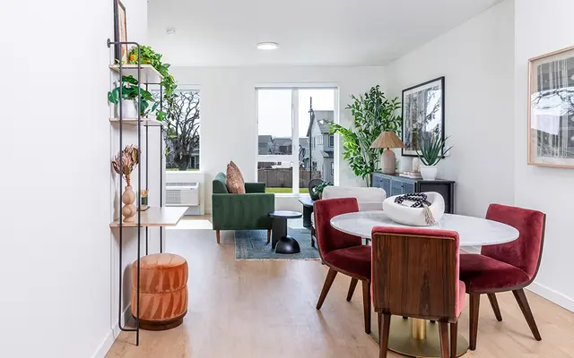 Modern Living Room and Dining Area A modern living room and dining area featuring a round dining table with upholstered chairs, a green sofa, and various indoor plants. Large windows allow natural light to enter.