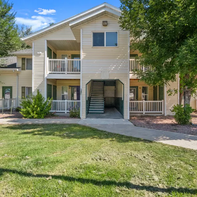 Exterior view of a two-story apartment building with balconies, surrounded by mature trees and landscaping. The building has a welcoming entryway with stairs leading to the upper floor, providing a comfortable and inviting residential atmosphere.