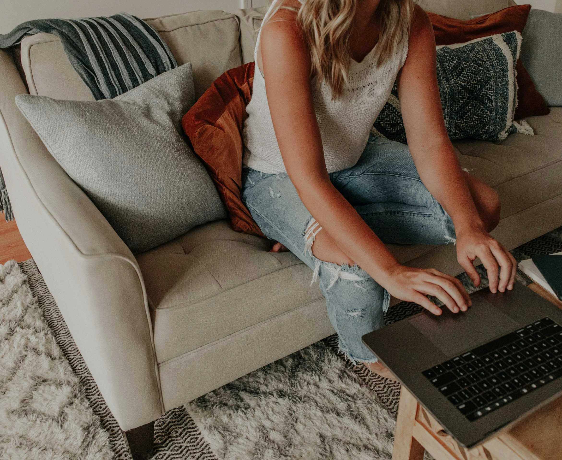 A woman sitting on a couch, working on a laptop. She is wearing a white top and distressed jeans, with cushions and a throw blanket around her.