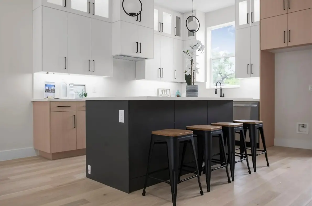 A modern kitchen featuring a large central island with black cabinetry and wooden stools. The kitchen has white cabinets and light wood flooring, with a window allowing natural light to enter. Decorative elements include an orchid plant and minimalist light fixtures.
