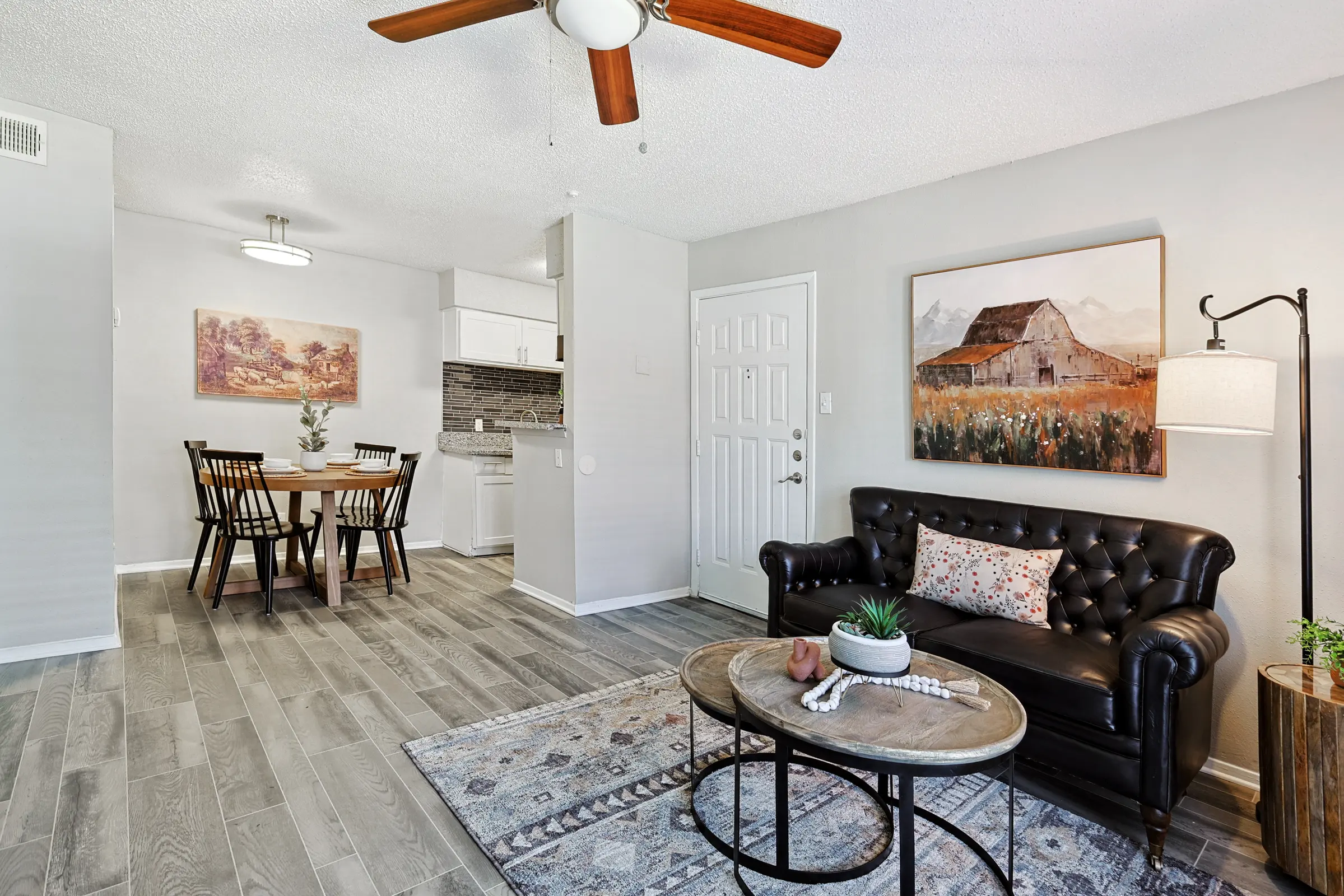 A stylish modern living room with a black leather sofa, a round coffee table, and wooden dining set visible in the background. The room features light grey walls, wooden flooring, and a ceiling fan. A painting of a barn is on the wall beside the entrance.