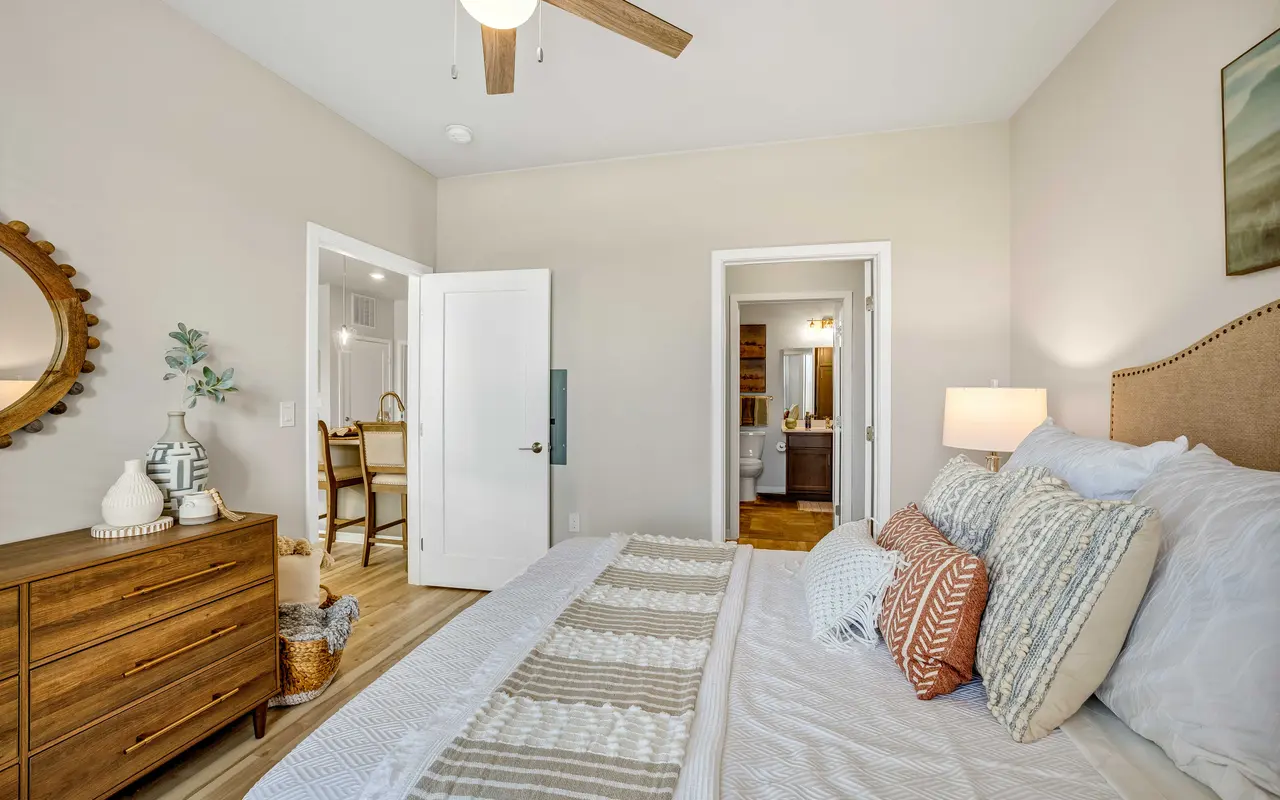 A modern bedroom featuring a neatly made bed with decorative pillows, a wooden dresser, a round mirror, and a ceiling fan. There are open doors leading to another room, and natural light fills the space.