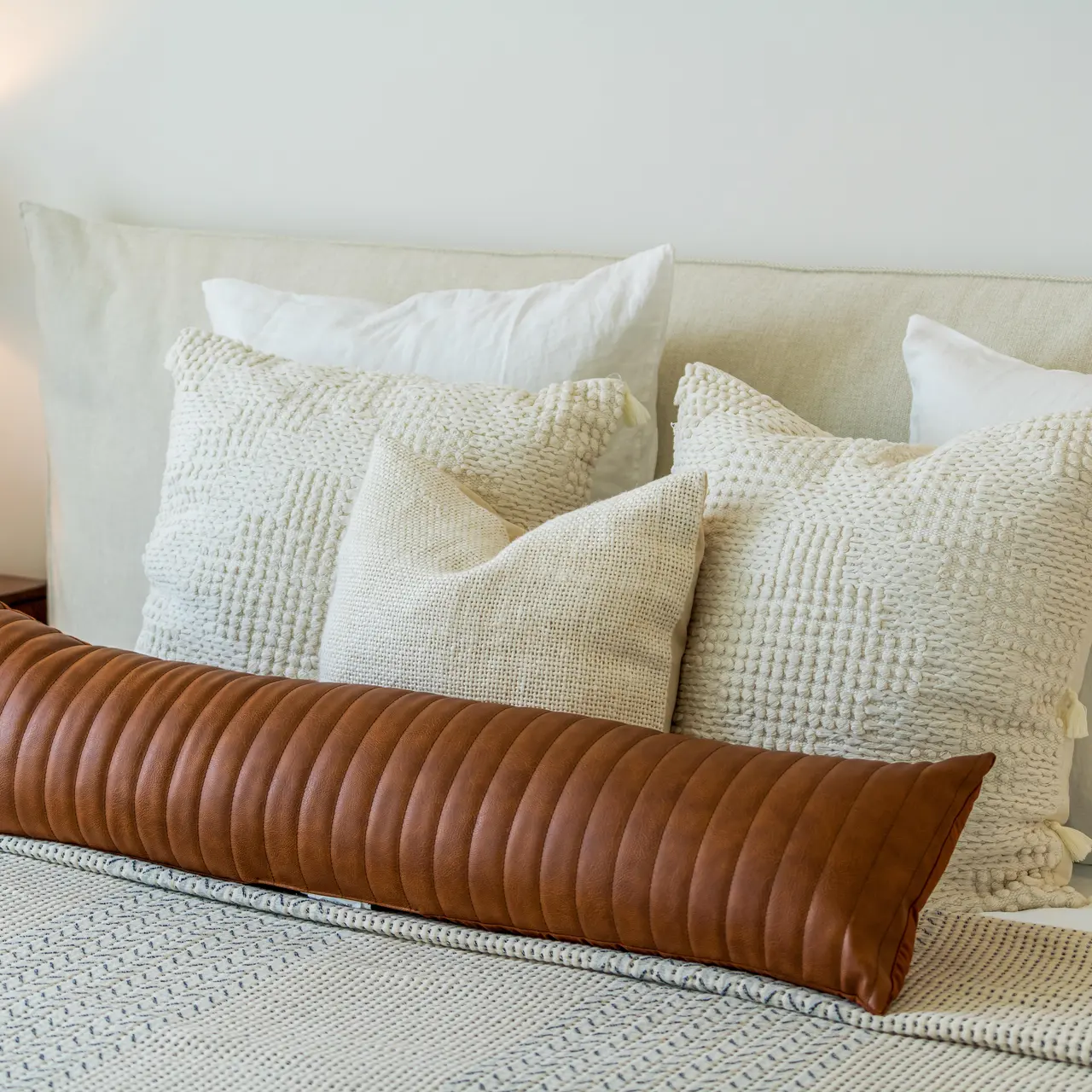 A well-decorated bed with various pillows including a long brown leather pillow and several textured white pillows against a light-colored wall.