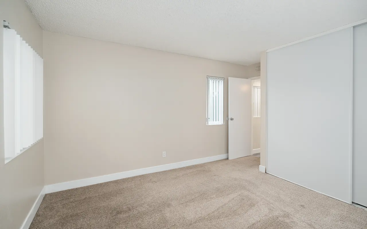 A spacious empty room with light beige walls and carpet. One wall features vertical blinds over two windows. A door on the right leads to another area, and there is a closet on the far end.