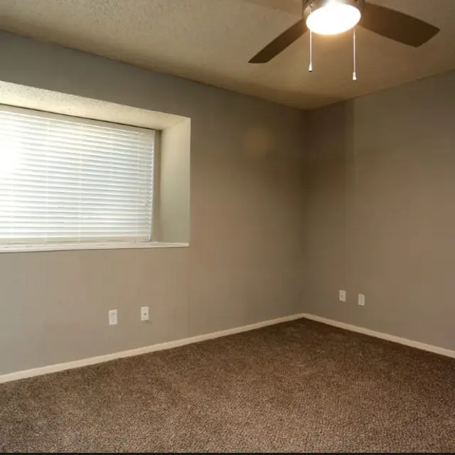An empty room with a ceiling fan and a large window featuring blinds. The walls are painted light gray, and the floor is carpeted in a light tan color.