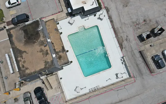 An aerial view of a pool surrounded by a concrete area with parked cars nearby and buildings in the background.