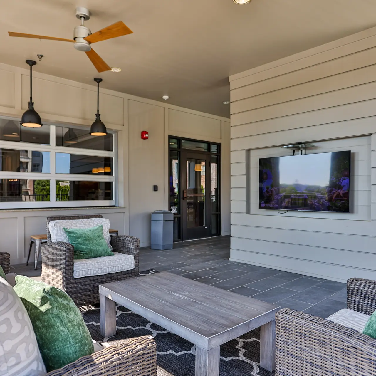 Outdoor Lounge Area with TV and Seating An outdoor lounge area featuring comfortable seating with decorative pillows, a table, and a wall-mounted television. The space is well-lit with ceiling fans and pendant lights.