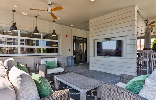 Outdoor Lounge Area with TV and Seating An outdoor lounge area featuring comfortable seating with decorative pillows, a table, and a wall-mounted television. The space is well-lit with ceiling fans and pendant lights.