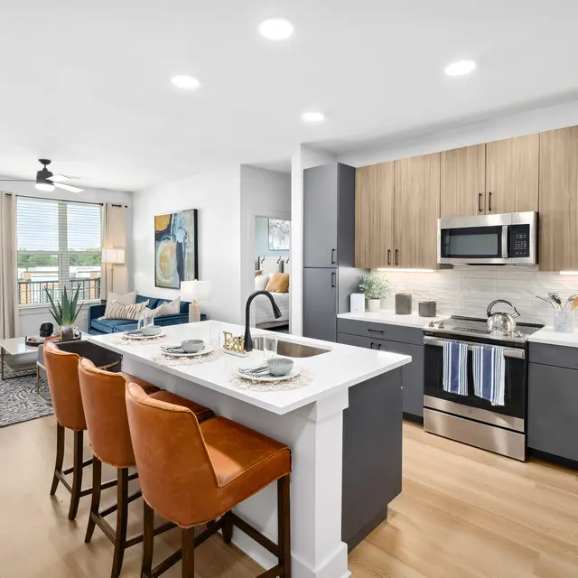 A modern apartment kitchen and living area featuring a sleek design with wooden cabinets, stainless steel appliances, and a breakfast bar with orange stools. Large windows provide natural light and a view of the outdoors.