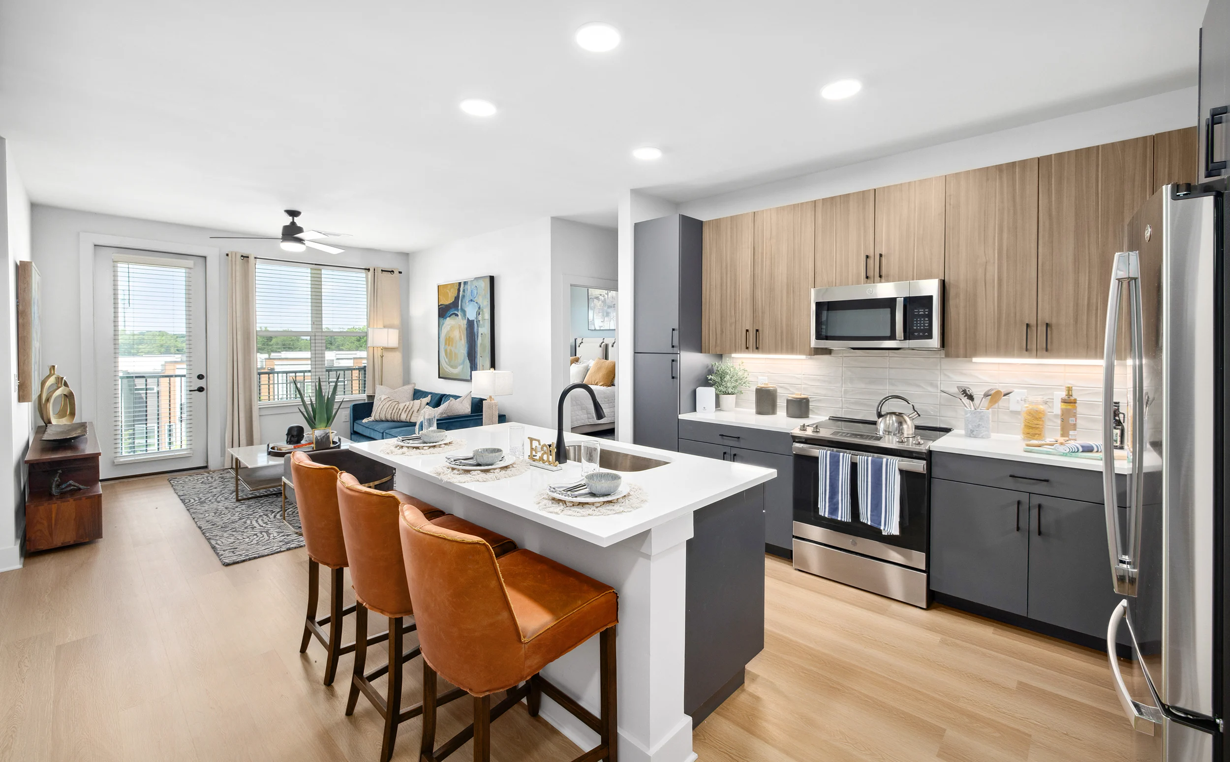 Contemporary Apartment Kitchen and Living Room A modern apartment kitchen and living area featuring a sleek design with wooden cabinets, stainless steel appliances, and a breakfast bar with orange stools. Large windows provide natural light and a view of the outdoors.