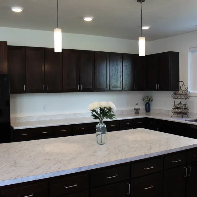Stylish Modern Kitchen Interior A modern kitchen featuring dark wooden cabinets, a large marble countertop island, and a black refrigerator. The kitchen has pendant lighting, a sink with a window, and decorative items on the shelves.